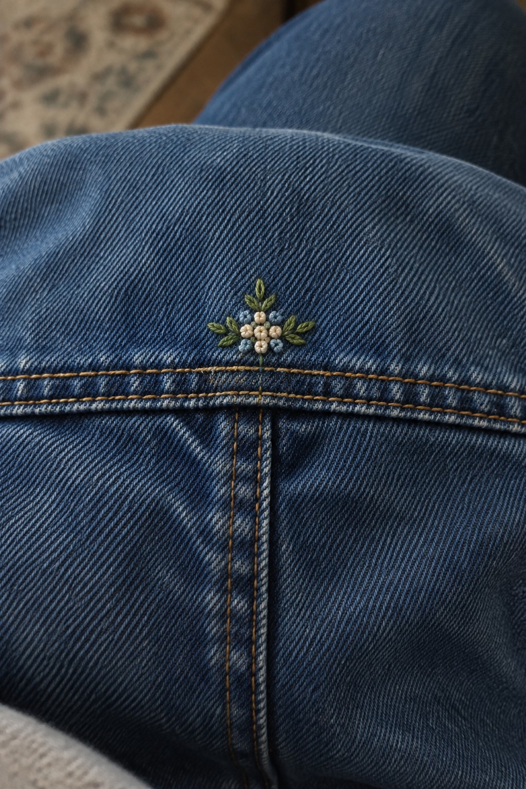 A small embroidered cluster of cream and pale blue flowers with green leaves sits above a seam on blue denim.
