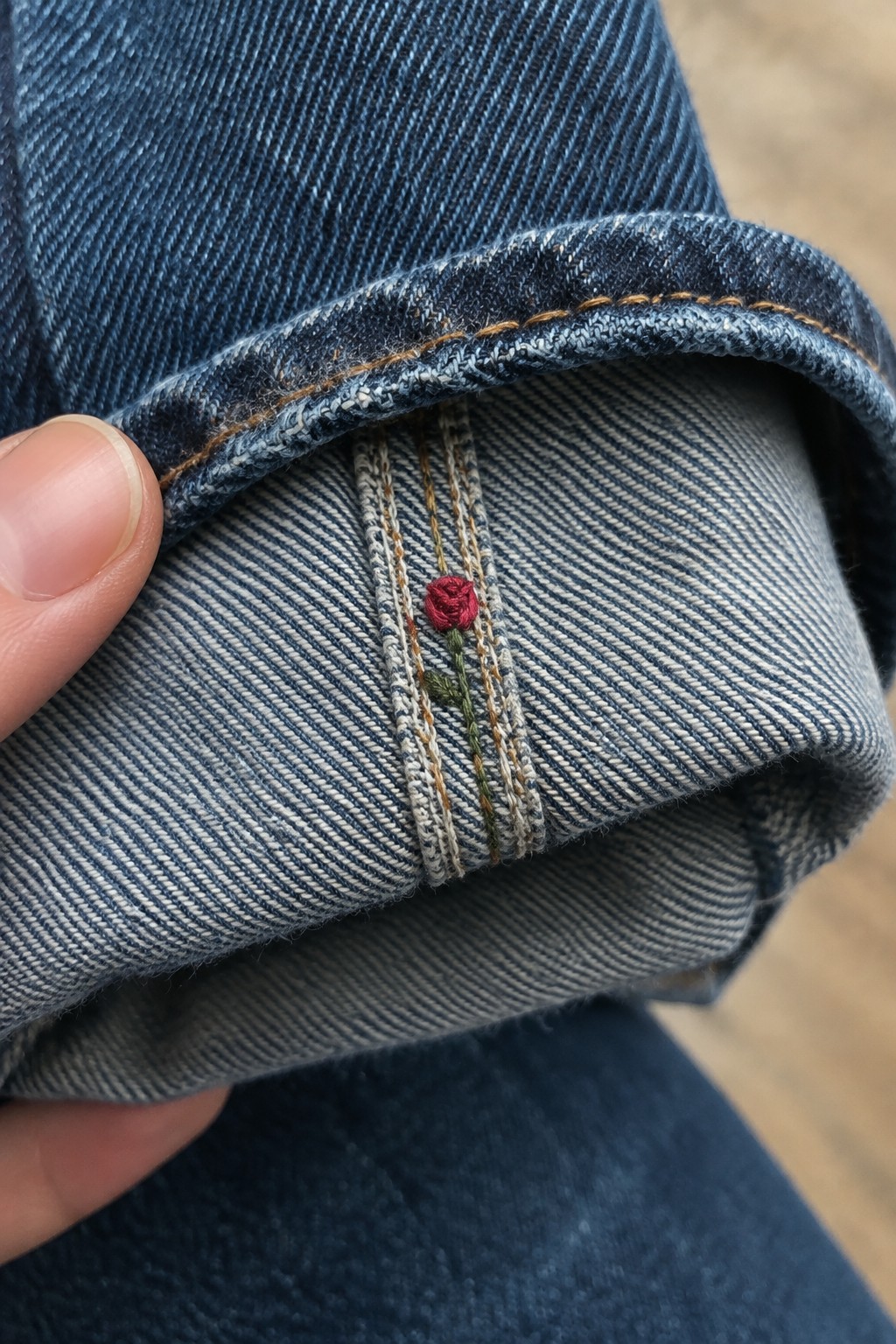 A small red rose with a green stem embroidered beside the seam on the turned-up cuff of blue jeans.