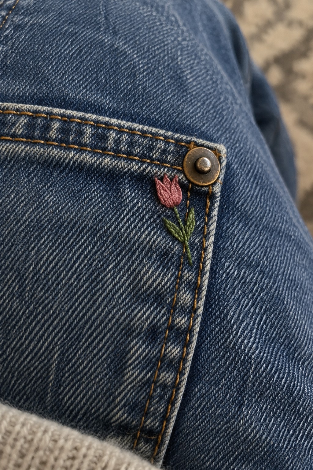 Close-up of a small pink tulip embroidered beside the rivet on a blue jeans pocket.