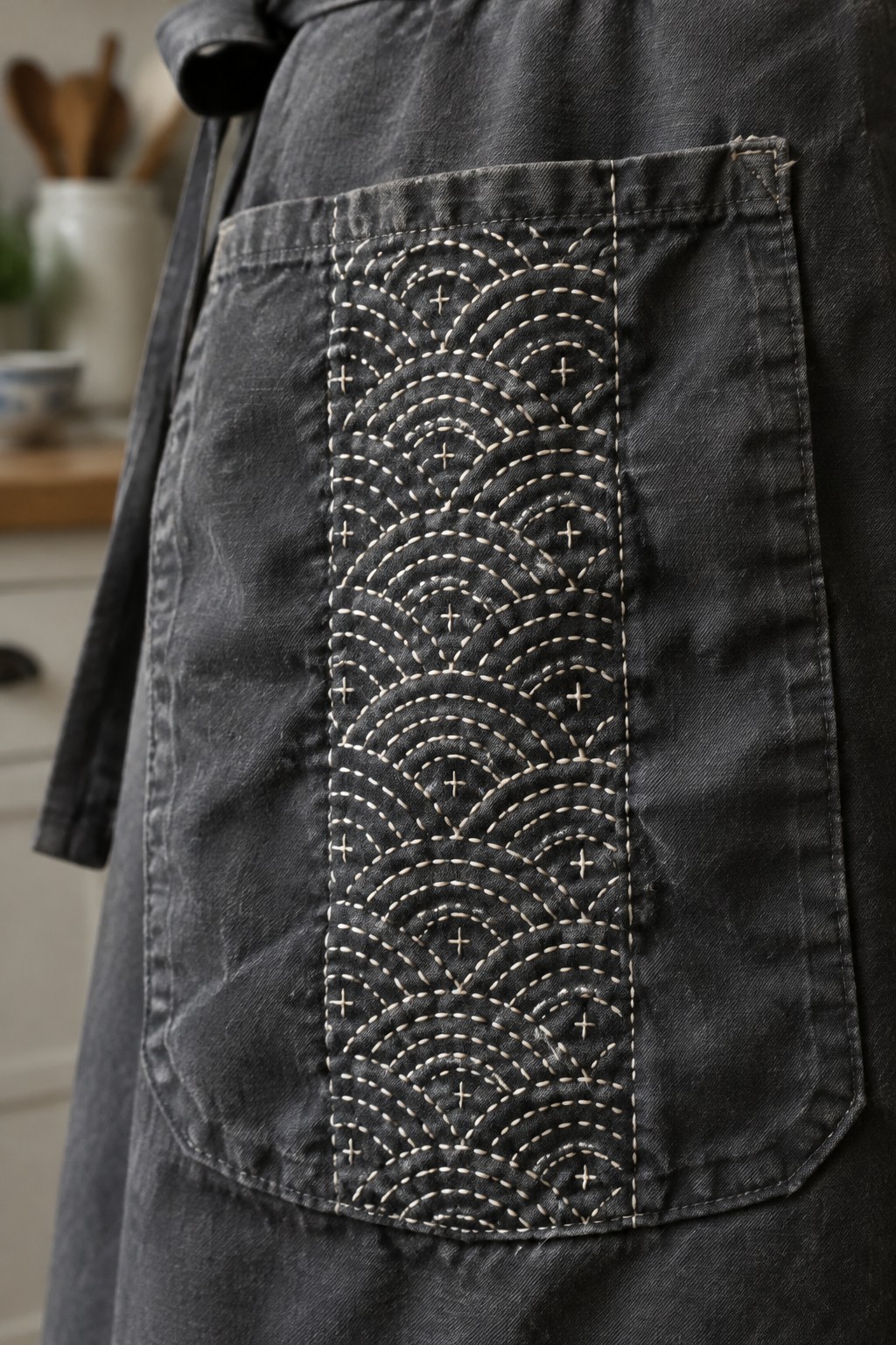 A dark denim apron pocket decorated with a vertical panel of cream repeating wave embroidery.