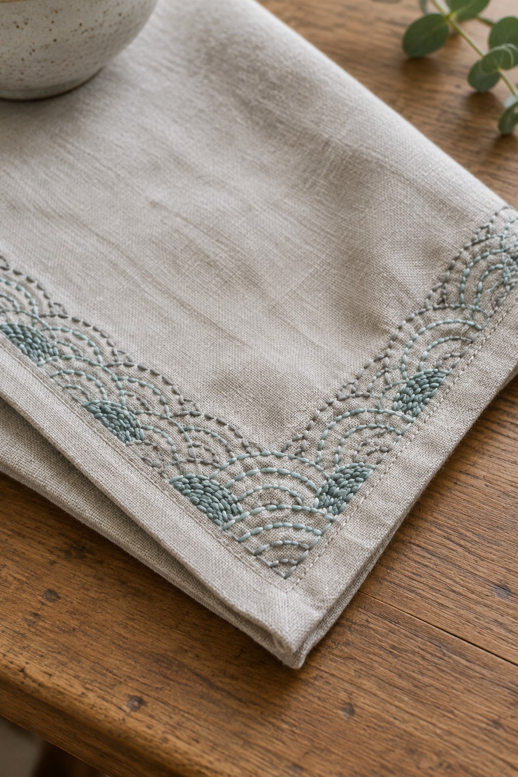 A folded beige linen runner with a blue-green and gray embroidered wave border rests on a wooden table.