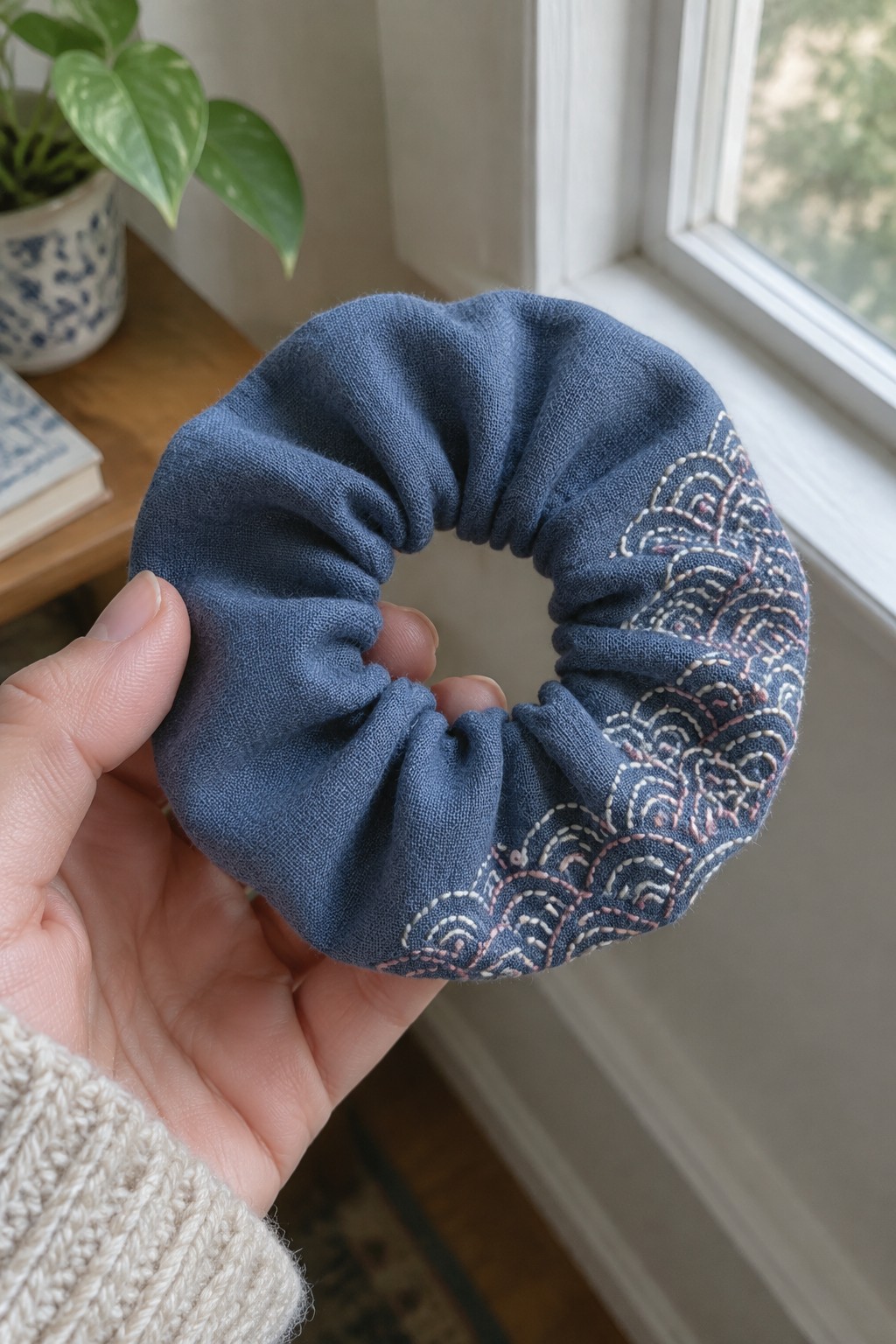 A hand holds a blue fabric scrunchie embroidered with pale repeating wave patterns along one side.