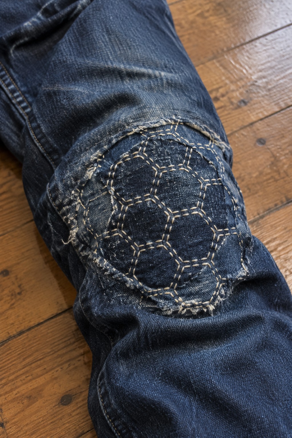 A round dark denim patch with cream geometric stitching repairs the knee of blue jeans.