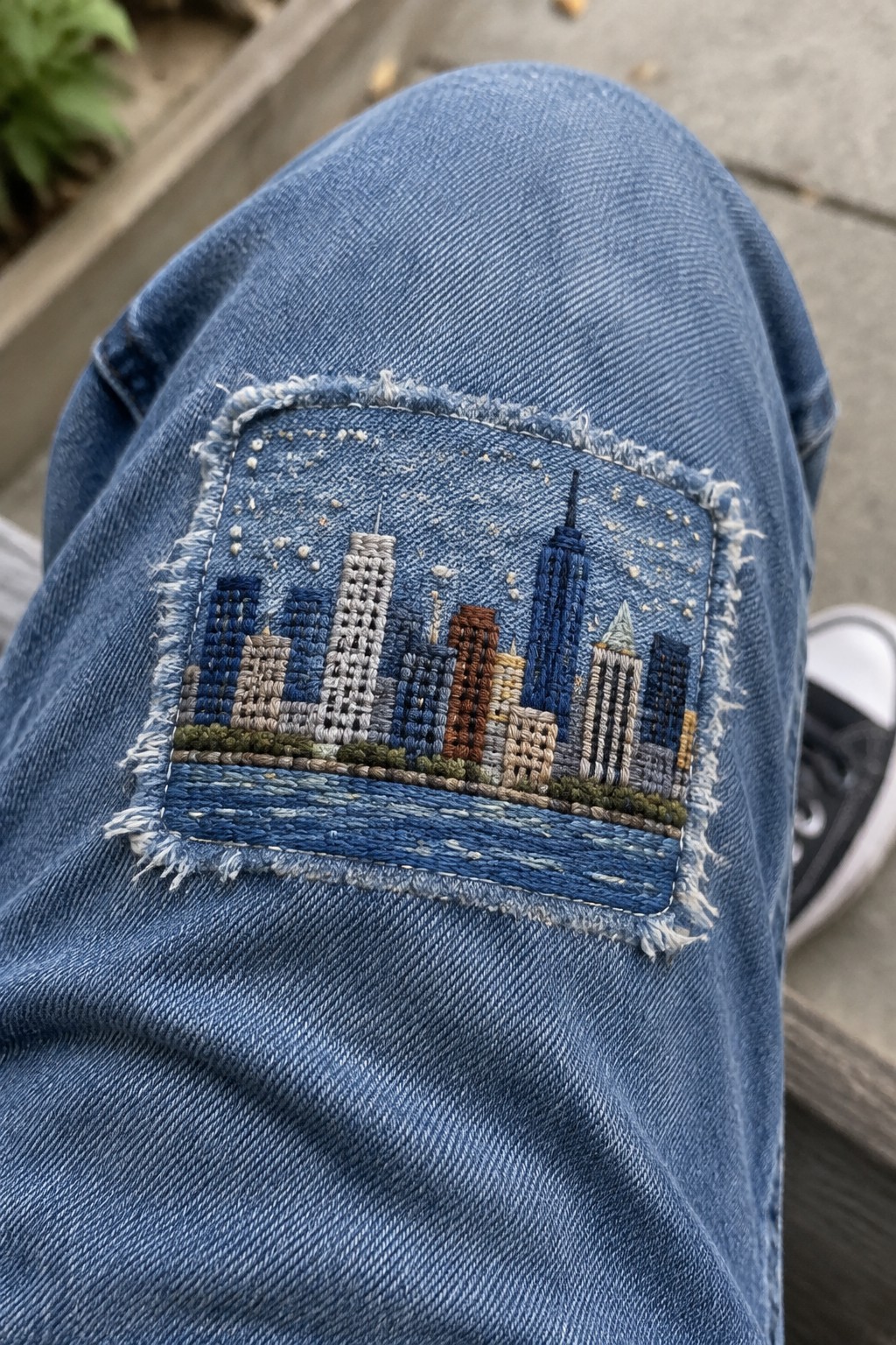 A rectangular embroidered city skyline and water patch is stitched onto the knee of blue jeans.