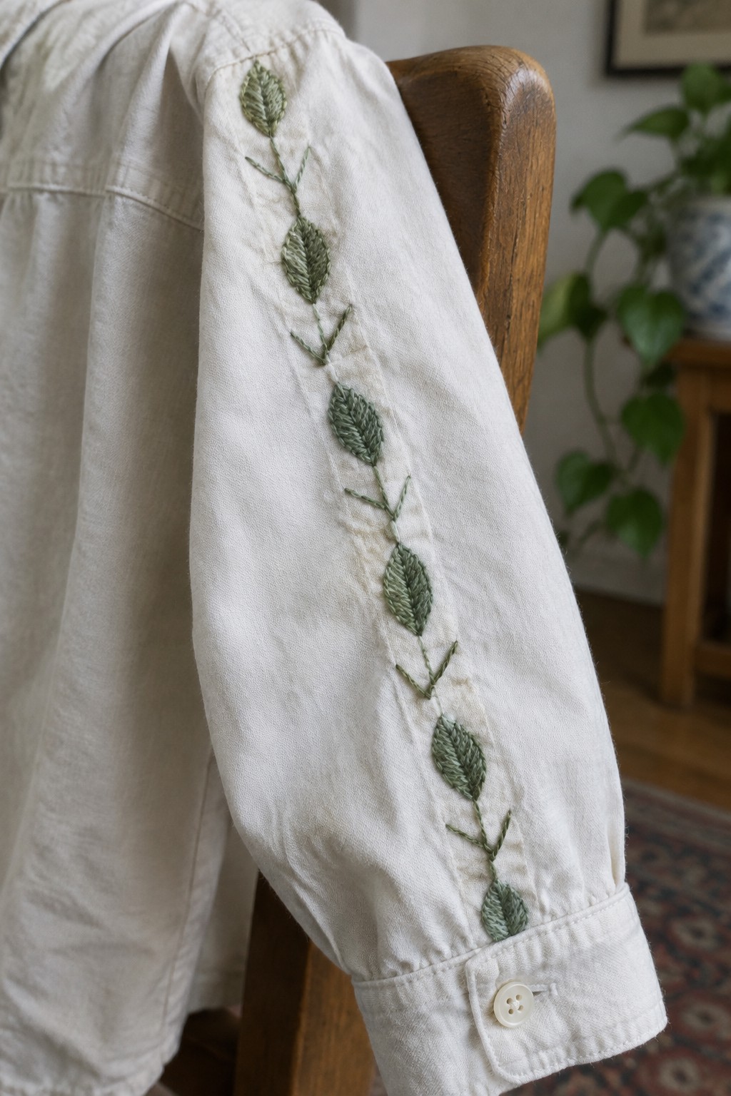 A white shirt sleeve with a green leafy vine embroidered down its length, draped over a wooden chair.