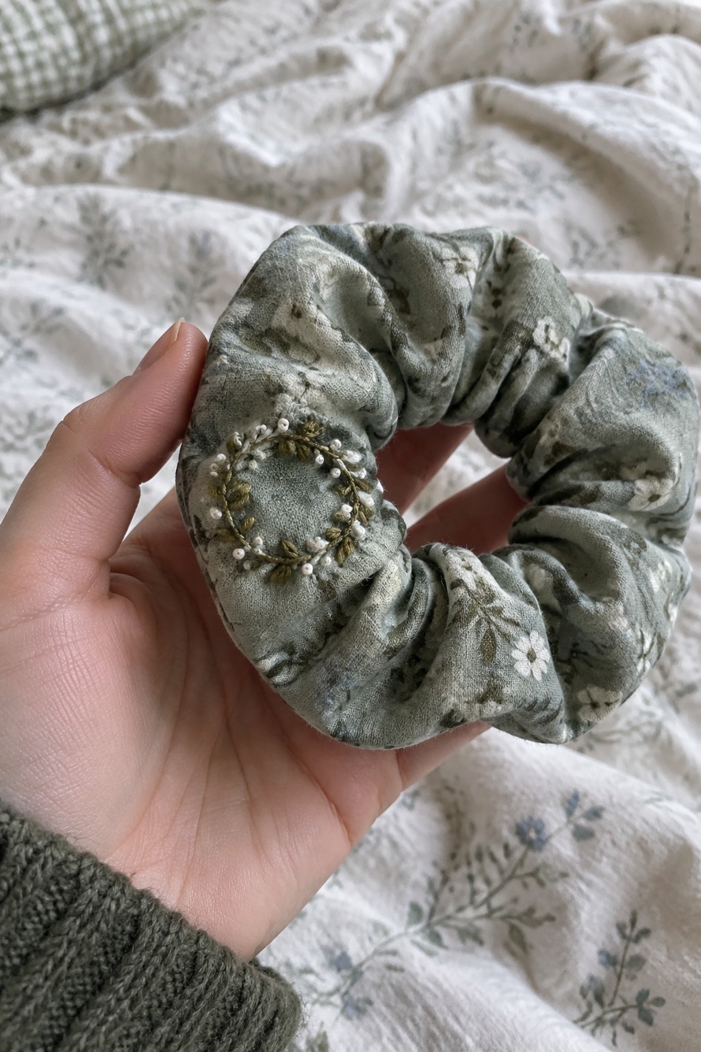 A hand holds a gathered green floral fabric scrunchie decorated with a small embroidered wreath of leaves and white accents.
