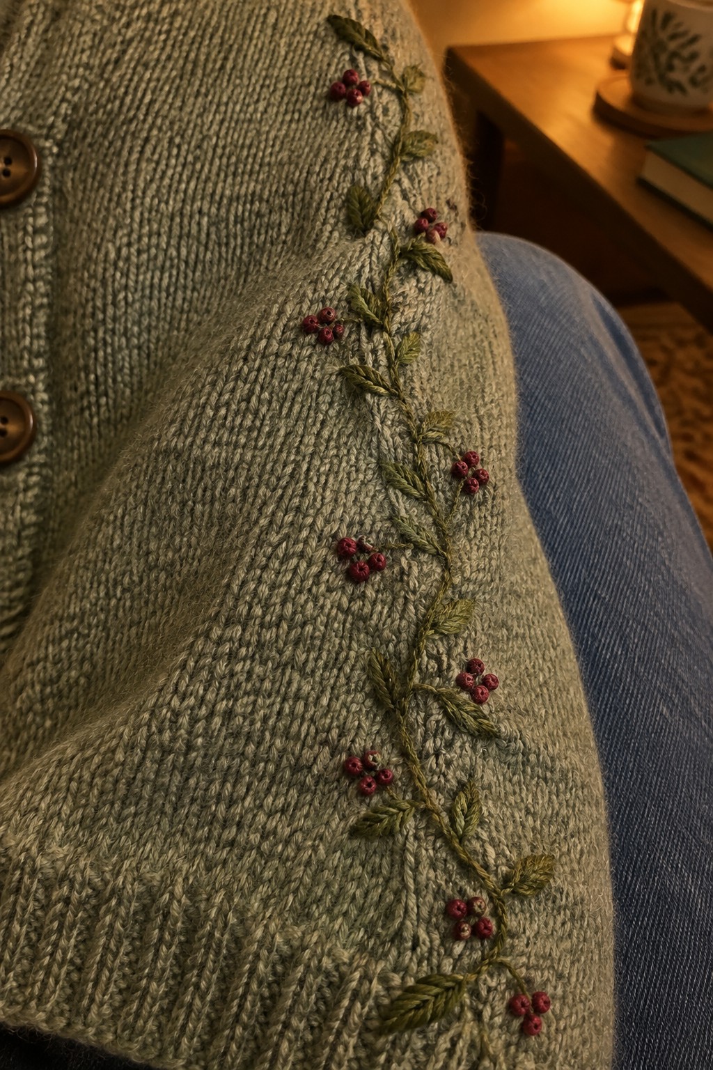 Green leafy vine embroidery with red berry clusters runs down the edge of a gray knit cardigan beside its buttons.