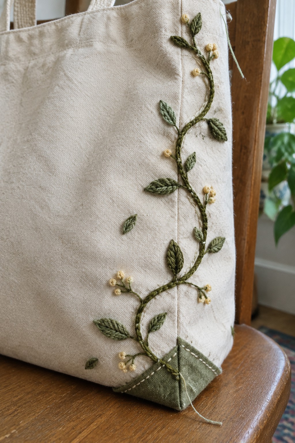 A cream canvas tote with a green embroidered vine and small cream buds beside its side seam.