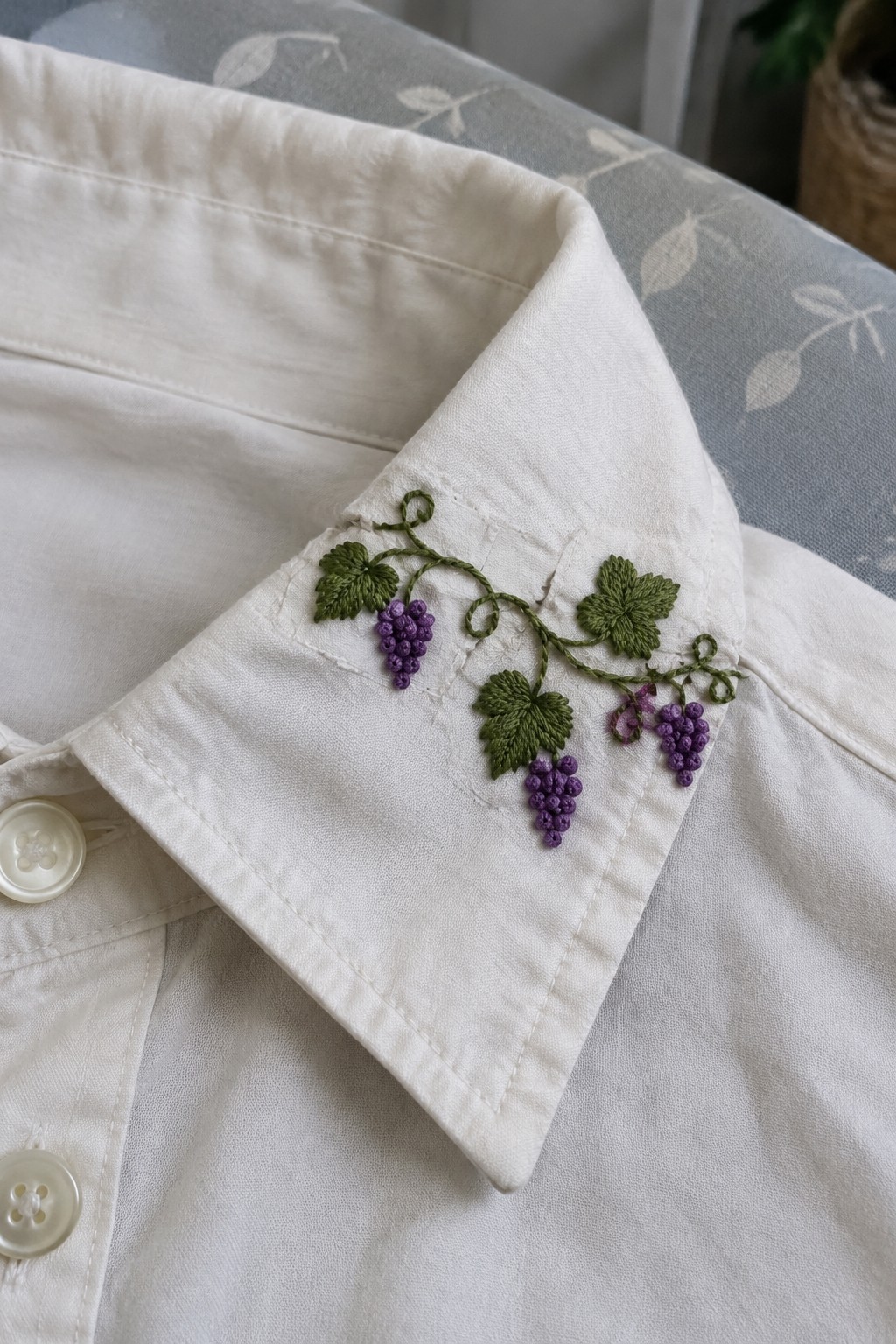 A white shirt cuff embroidered with green grape leaves and stems and three clusters of purple grapes.