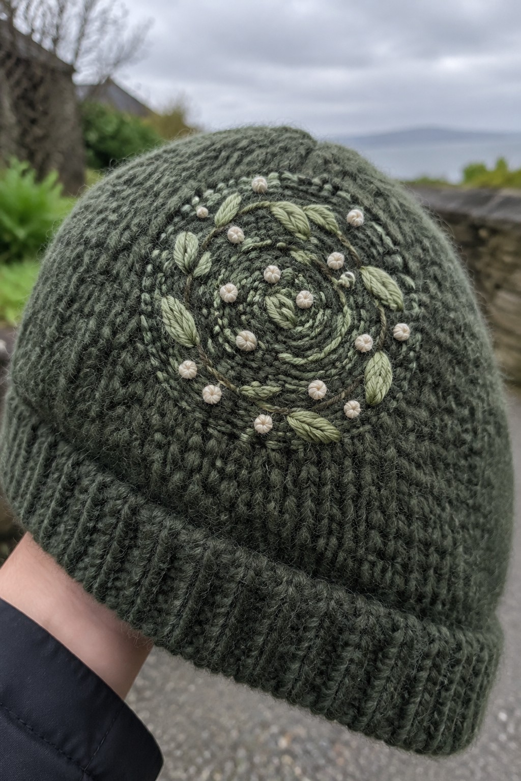Dark green knitted beanie with a spiral of embroidered light green leaves and cream flowers near the crown.