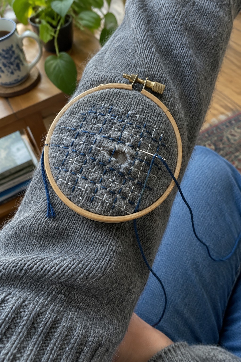 Blue and white crosshatched stitches mend a gray sweater elbow held in an embroidery hoop.