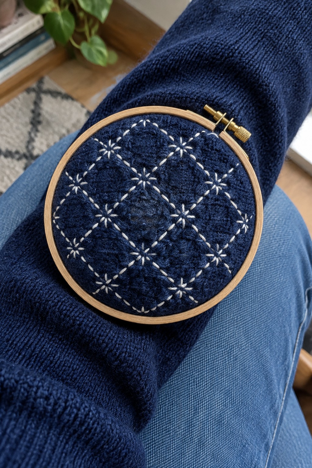White geometric stitching forms a repeating pattern on a dark blue knitted sweater inside an embroidery hoop.