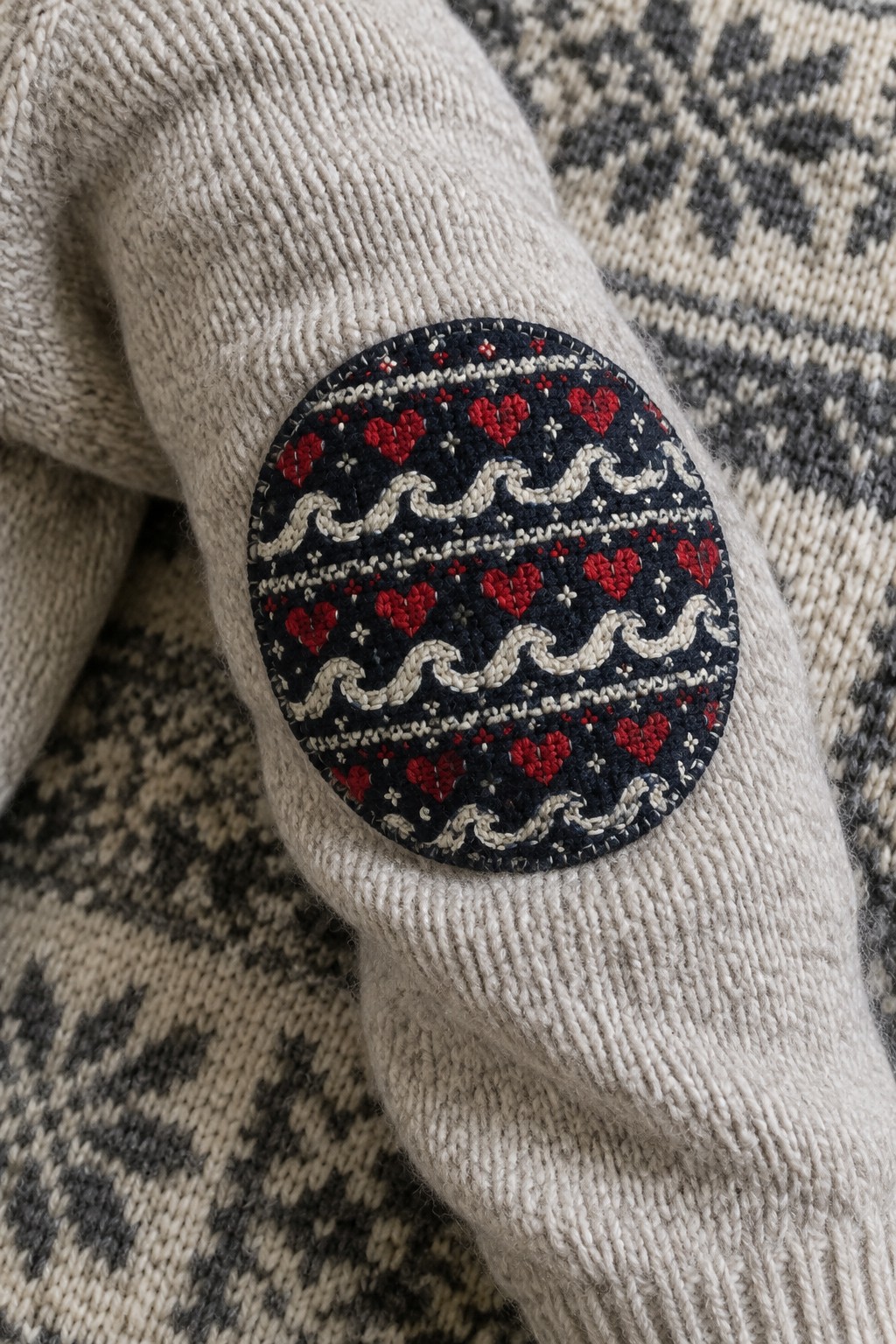 A navy oval patch embroidered with red hearts and cream wave bands covers the elbow of a light knitted sweater.