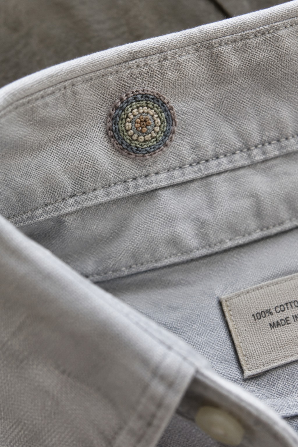 Close-up of a gray shirt placket with a small circular, multicolored embroidery motif and a fabric label.