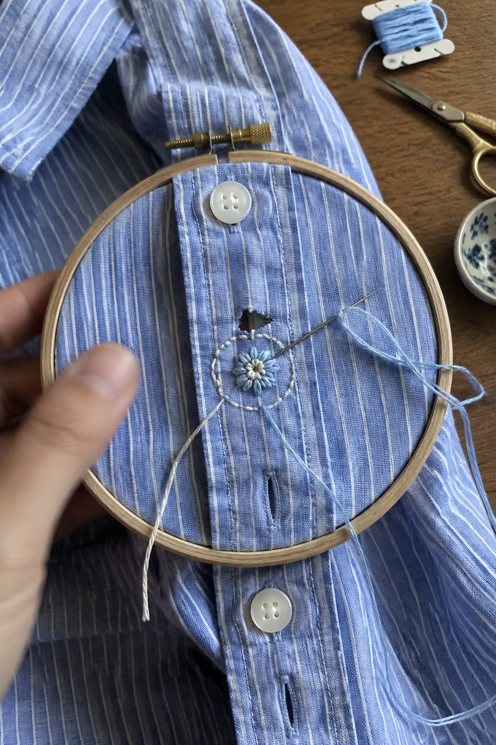 Blue embroidered flower and stitched circle cover a hole in a striped shirt placket inside an embroidery hoop.