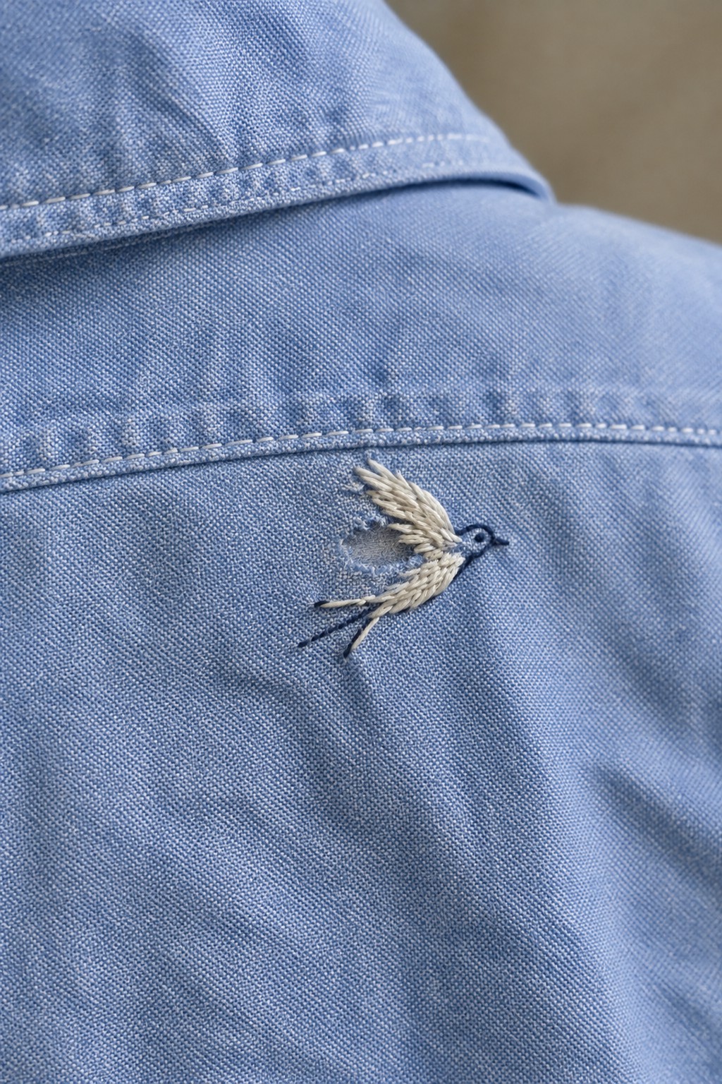 A small cream-and-dark-blue embroidered bird covers a hole in a blue shirt below a curved seam.
