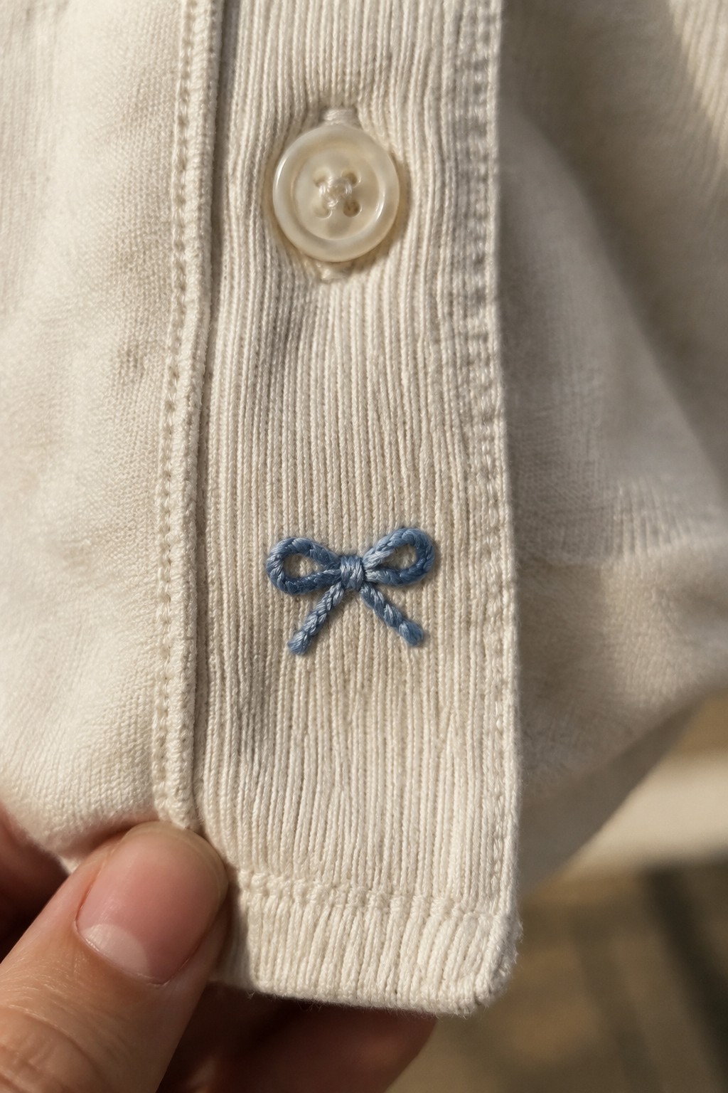 A small blue embroidered bow sits on the ribbed placket of a white shirt below a button.
