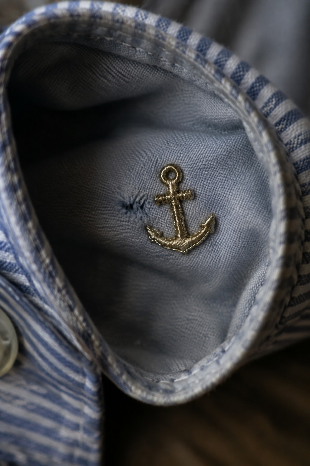 A small gold embroidered anchor beside a hole in the blue-gray fabric inside a striped shirt collar.