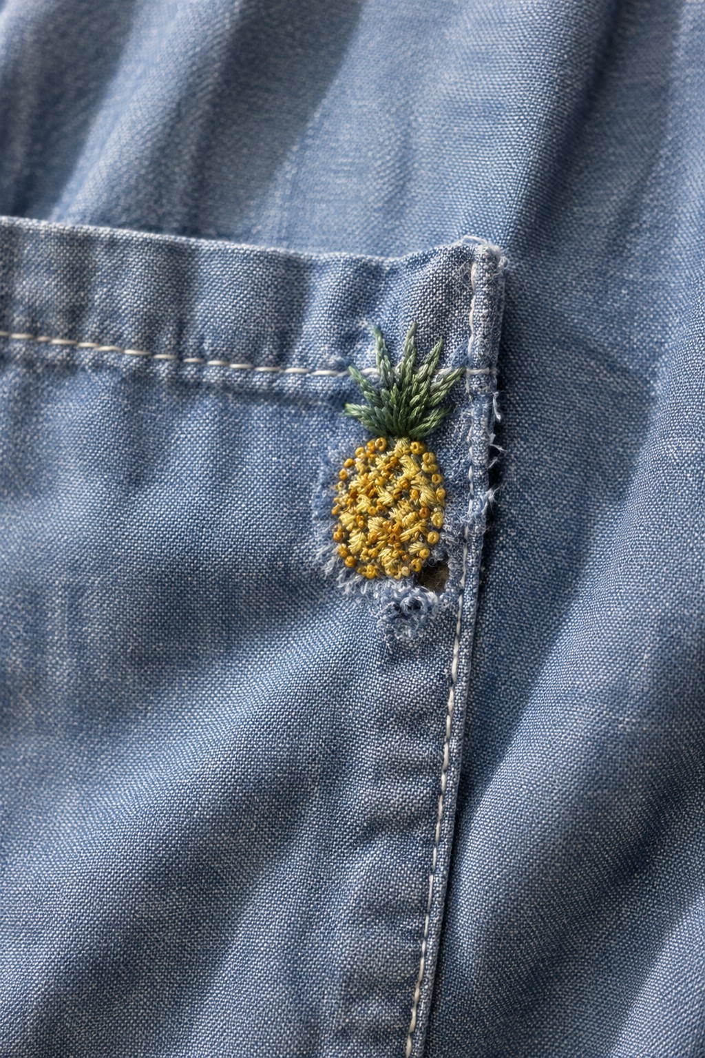 A small yellow and green embroidered pineapple covers a frayed hole beside the corner of a blue denim pocket.
