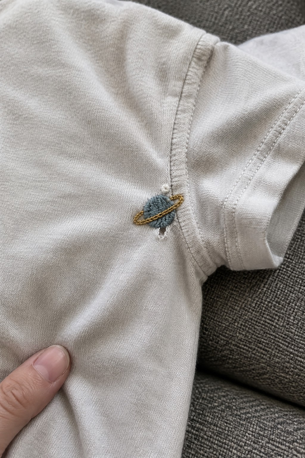 A small blue-green planet with a gold ring embroidered beside the seam of a light-colored shirt.